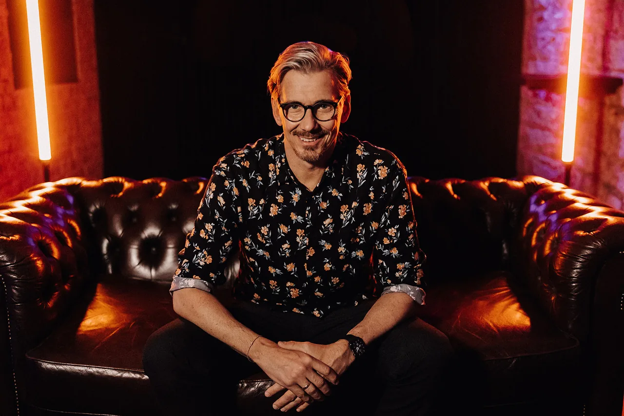 Alex Maus sitzt auf einem Ledersofa im Neonlicht