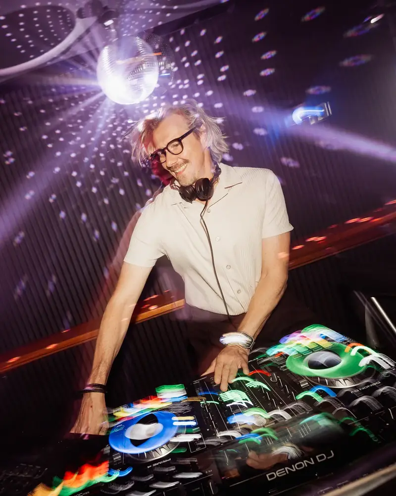 Alex lacht am DJ Pult unter der Discokugel